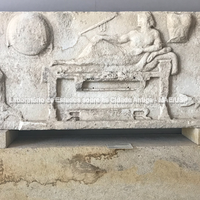 Slab (A758) Archaeological Museum of Paros.
A cena mostra o próprio Archilochos reclinado em um sofá, sua esposa sentada à esquerda e um garoto à direita, que oferece vinho ao poeta. Seus braços estão pendurados; na parede: peitoral, escudo, aljava e lira -, foram encontrados no chão da igreja de Ekatontapyliani. As duas lajes prováveis provêm do chamado Archilocheion
Archilochus foi homenageado em Paros como poeta e guerreiro no final do século VI, e se gabou de que ele era o servo de Lorde Enyalios e entendeu que o belo presente das musas encontra seu reconhecimento em um monumento, que o exibe banquetes no presença dos vivos e recebimento das ofertas pelas quais eles o honravam como guerreiro e como poeta.
